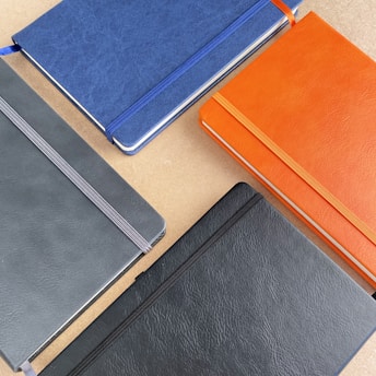 Four hardcover notebooks with elastic bands are placed on a light brown surface. The notebooks are in different colors: blue, orange, dark gray, and black. Each notebook has a fabric bookmark ribbon matching the color of the elastic band.