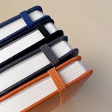 A set of four stacked journals with hardcovers and elastic bands securing them. The colors of the covers and bands include blue, black, gray, and orange. The journals are arranged neatly from a close-up perspective.