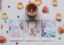 Three tarot cards are placed on a flat surface surrounded by scattered flower petals and a lit candle. The cards are titled 'The Age of Light', 'Dance with Life', and 'Starseed', each with illustrated imagery and accompanying subtitles. The candle is centrally located above the cards, casting a warm glow.