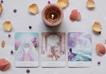 Three tarot cards are placed on a flat surface surrounded by scattered flower petals and a lit candle. The cards are titled 'The Age of Light', 'Dance with Life', and 'Starseed', each with illustrated imagery and accompanying subtitles. The candle is centrally located above the cards, casting a warm glow.