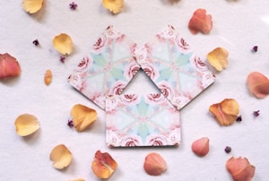 Three beautifully designed cards with intricate floral patterns featuring roses and geometric shapes are placed on a soft, white surface. They are surrounded by scattered petals in shades of pink and yellow, along with a few tiny purple buds.