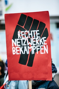 A protest sign featuring a stylized black fist on a red background with bold white lettering that reads 'RECHTE NETZWERKE BEKÄMPFEN'. The image captures a scene from a demonstration with people in the background.