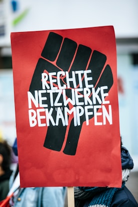 A protest sign featuring a stylized black fist on a red background with bold white lettering that reads 'RECHTE NETZWERKE BEKÄMPFEN'. The image captures a scene from a demonstration with people in the background.