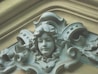 A detailed and ornate architectural sculpture of a serene-faced female figure, possibly made of stone or plaster, surrounded by intricate floral and scrollwork patterns. The figure has wavy hair and is integrated into a decorative element, possibly on a building facade.