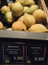 A basket filled with fresh yellow and green lemons with price tags labeled in German. The labels indicate the lemons are organic and display prices per kilogram or per piece.