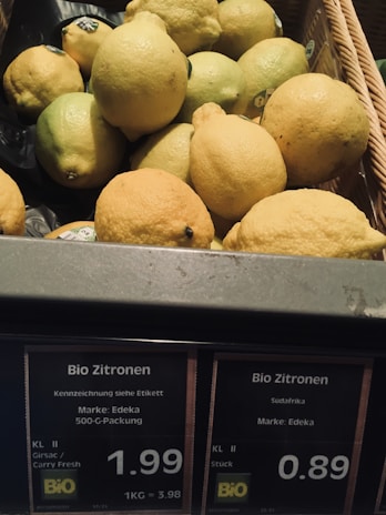 A basket filled with fresh yellow and green lemons with price tags labeled in German. The labels indicate the lemons are organic and display prices per kilogram or per piece.