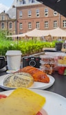 An outdoor breakfast spread on a table features a variety of pastries, including a croissant and a poppy seed bun, alongside slices of cheese. There are several containers with snacks, a disposable coffee cup, and two cameras in the background. The scene is set in front of a historic building with a garden and tents in the background.