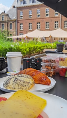 An outdoor breakfast spread on a table features a variety of pastries, including a croissant and a poppy seed bun, alongside slices of cheese. There are several containers with snacks, a disposable coffee cup, and two cameras in the background. The scene is set in front of a historic building with a garden and tents in the background.