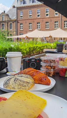 An outdoor breakfast spread on a table features a variety of pastries, including a croissant and a poppy seed bun, alongside slices of cheese. There are several containers with snacks, a disposable coffee cup, and two cameras in the background. The scene is set in front of a historic building with a garden and tents in the background.