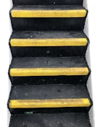 A set of black stairs features bright yellow anti-slip strips on the edge of each step, with white walls on either side, creating a stark contrast.