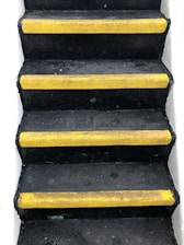 A set of black stairs features bright yellow anti-slip strips on the edge of each step, with white walls on either side, creating a stark contrast.