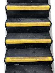 A set of black stairs features bright yellow anti-slip strips on the edge of each step, with white walls on either side, creating a stark contrast.
