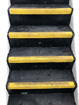 A set of black stairs features bright yellow anti-slip strips on the edge of each step, with white walls on either side, creating a stark contrast.