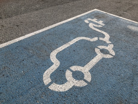 A blue parking space is marked with a white line drawing of an electric car and a charging plug, indicating a designated spot for electric vehicle charging.