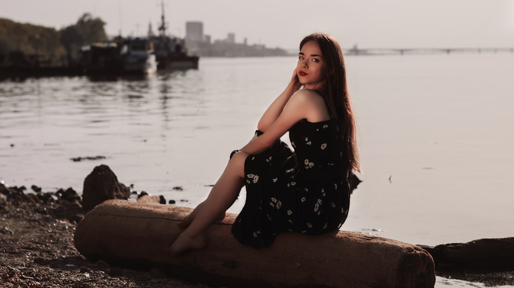 woman in black and white floral dress sitting on brown wooden log near body of water