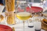 A glass of white wine is placed on a table set for dining, surrounded by red plates and clear glasses. A bundle of dry pasta tied with string is visible in the background, along with various glass containers. The setting appears to be outdoors, with soft natural lighting.
