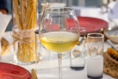 A glass of white wine is placed on a table set for dining, surrounded by red plates and clear glasses. A bundle of dry pasta tied with string is visible in the background, along with various glass containers. The setting appears to be outdoors, with soft natural lighting.