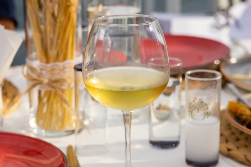 A glass of white wine is placed on a table set for dining, surrounded by red plates and clear glasses. A bundle of dry pasta tied with string is visible in the background, along with various glass containers. The setting appears to be outdoors, with soft natural lighting.