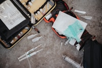 An assortment of medical supplies is scattered on a concrete floor, including syringes, vials, and medical bags filled with other equipment. The items appear to be part of an emergency medical kit or a mobile health unit.