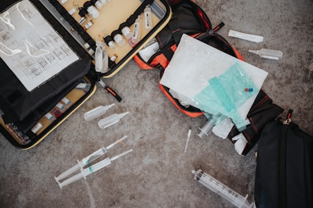An assortment of medical supplies is scattered on a concrete floor, including syringes, vials, and medical bags filled with other equipment. The items appear to be part of an emergency medical kit or a mobile health unit.