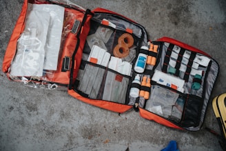 An open first aid kit is laid out on a concrete surface. It contains various medical supplies including bandages, bottles, and medical tools, organized neatly in mesh pockets. The kit is red with black trim.