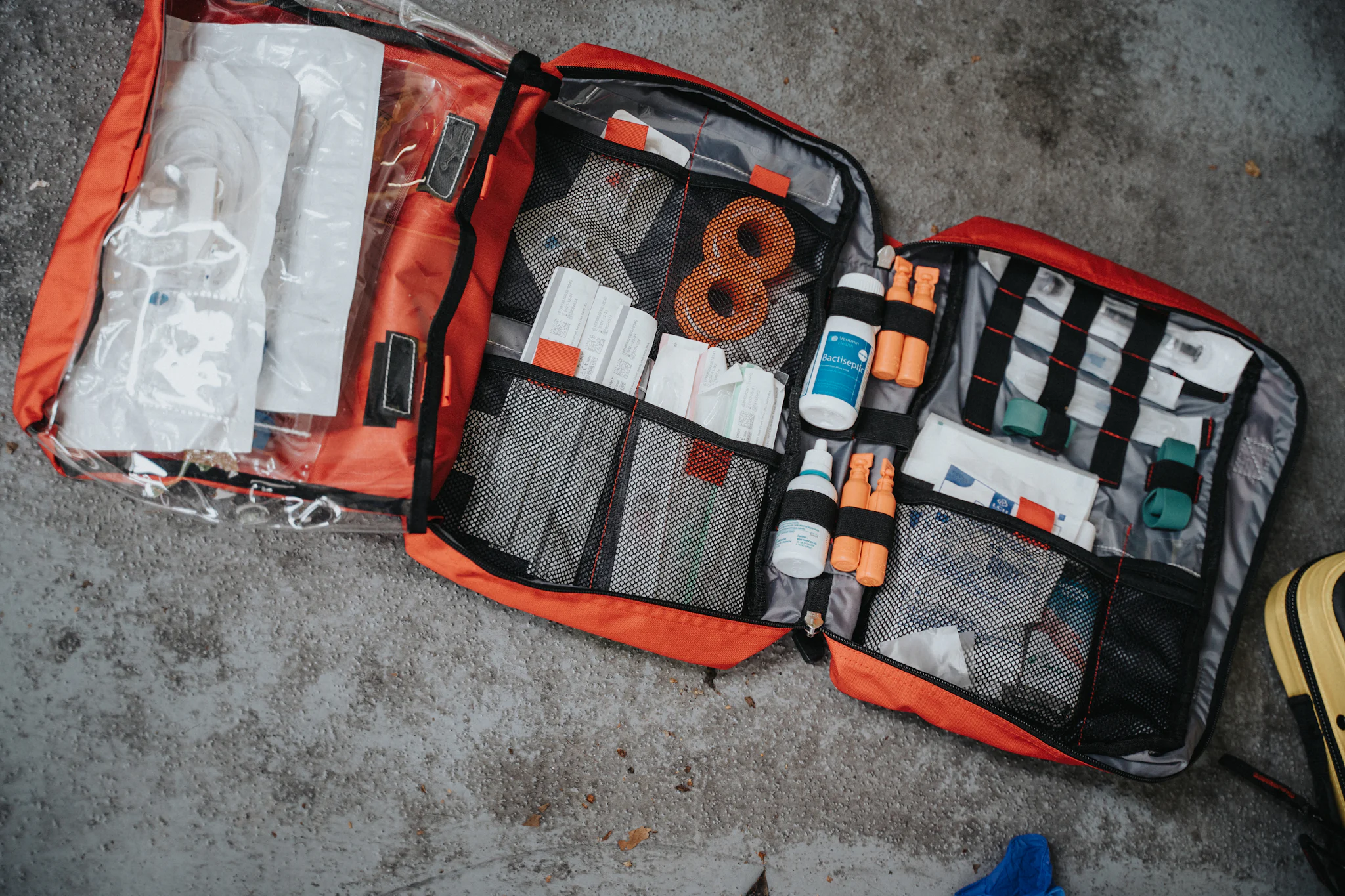 An open orange first aid bag on a concrete floor, its pockets filled with bandages, dressings and bottles