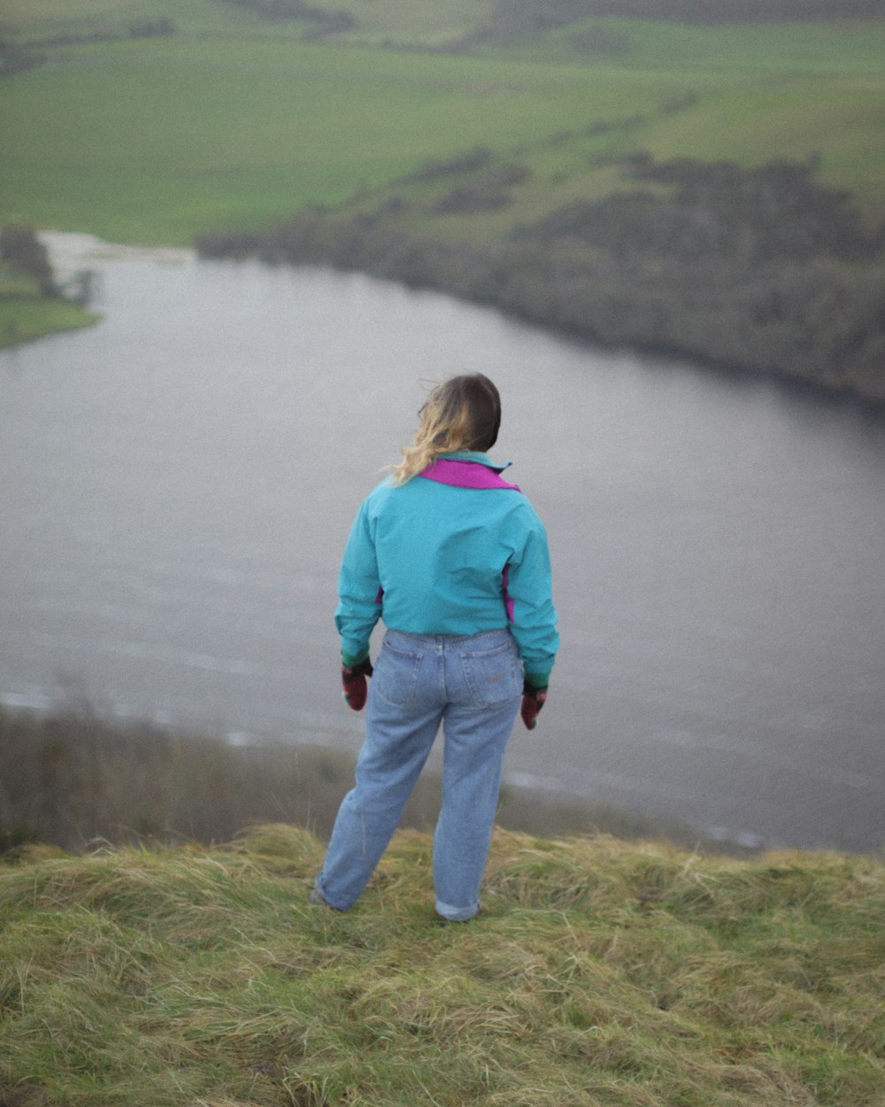 girl in blue jacket and blue denim jeans standing on green grass near lake during daytime