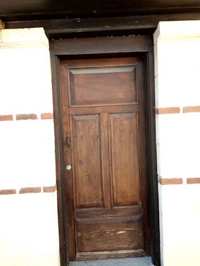 A wooden door with a natural finish set into a brick wall. The door features vertical panels and a rectangular window section at the top. The surrounding wall is made of light-colored bricks with some brown horizontal brick accents.