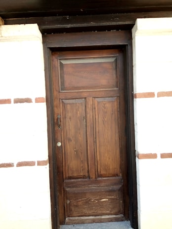A wooden door with a natural finish set into a brick wall. The door features vertical panels and a rectangular window section at the top. The surrounding wall is made of light-colored bricks with some brown horizontal brick accents.
