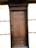 A wooden door with a natural finish set into a brick wall. The door features vertical panels and a rectangular window section at the top. The surrounding wall is made of light-colored bricks with some brown horizontal brick accents.