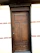 A wooden door with a natural finish set into a brick wall. The door features vertical panels and a rectangular window section at the top. The surrounding wall is made of light-colored bricks with some brown horizontal brick accents.