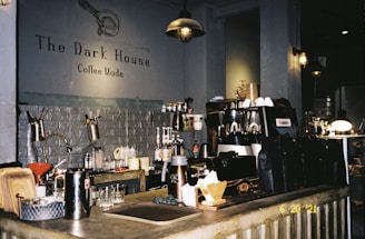 A cozy coffee shop interior with industrial lighting and a rustic counter. The counter holds various coffee-making equipment, cups, and coffee bags. The wall displays the shop name 'The Dark House' and 'Coffee Mode'. The atmosphere is warm and inviting.