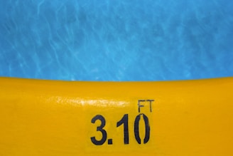 A bright yellow pool edge with the depth marking of 3.10 feet written in black. The blue pool water contrasts against the yellow edge, showing ripples and reflections.