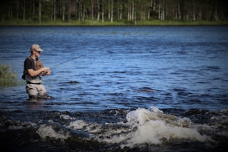 A person stands in a body of water, engaged in fly fishing. The water shows gentle ripples and small waves in the foreground, while the background features lush greenery and a forest under a clear blue sky.