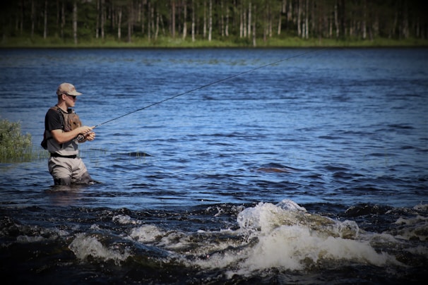 A person stands in a body of water, engaged in fly fishing. The water shows gentle ripples and small waves in the foreground, while the background features lush greenery and a forest under a clear blue sky.
