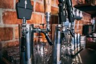Several beer taps are mounted on a shiny metallic column against a background of red brick walls. Glasses are placed beneath the taps, ready for use.