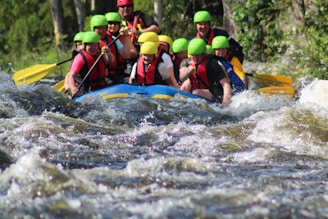 A group of people is white-water rafting down a river, wearing colorful helmets and life vests. The water is turbulent, with visible rapids and waves splashing around the raft. The participants appear focused and energetic as they navigate the fast-flowing water. Surrounding the river are lush green trees and vegetation.