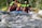 A group of people is white-water rafting down a river, wearing colorful helmets and life vests. The water is turbulent, with visible rapids and waves splashing around the raft. The participants appear focused and energetic as they navigate the fast-flowing water. Surrounding the river are lush green trees and vegetation.