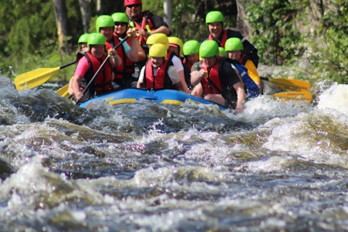 A group of people is white-water rafting down a river, wearing colorful helmets and life vests. The water is turbulent, with visible rapids and waves splashing around the raft. The participants appear focused and energetic as they navigate the fast-flowing water. Surrounding the river are lush green trees and vegetation.