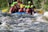 A group of people is white-water rafting down a river, wearing colorful helmets and life vests. The water is turbulent, with visible rapids and waves splashing around the raft. The participants appear focused and energetic as they navigate the fast-flowing water. Surrounding the river are lush green trees and vegetation.