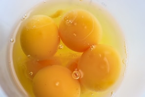 Four raw egg yolks floating in egg white inside a white bowl. Small bubbles are present on the surface of the egg whites.