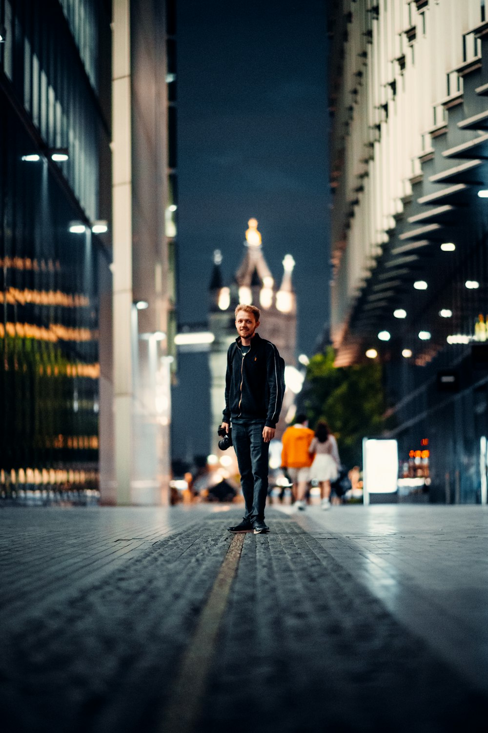 homem em jaqueta preta e calças pretas em pé na calçada durante a noite