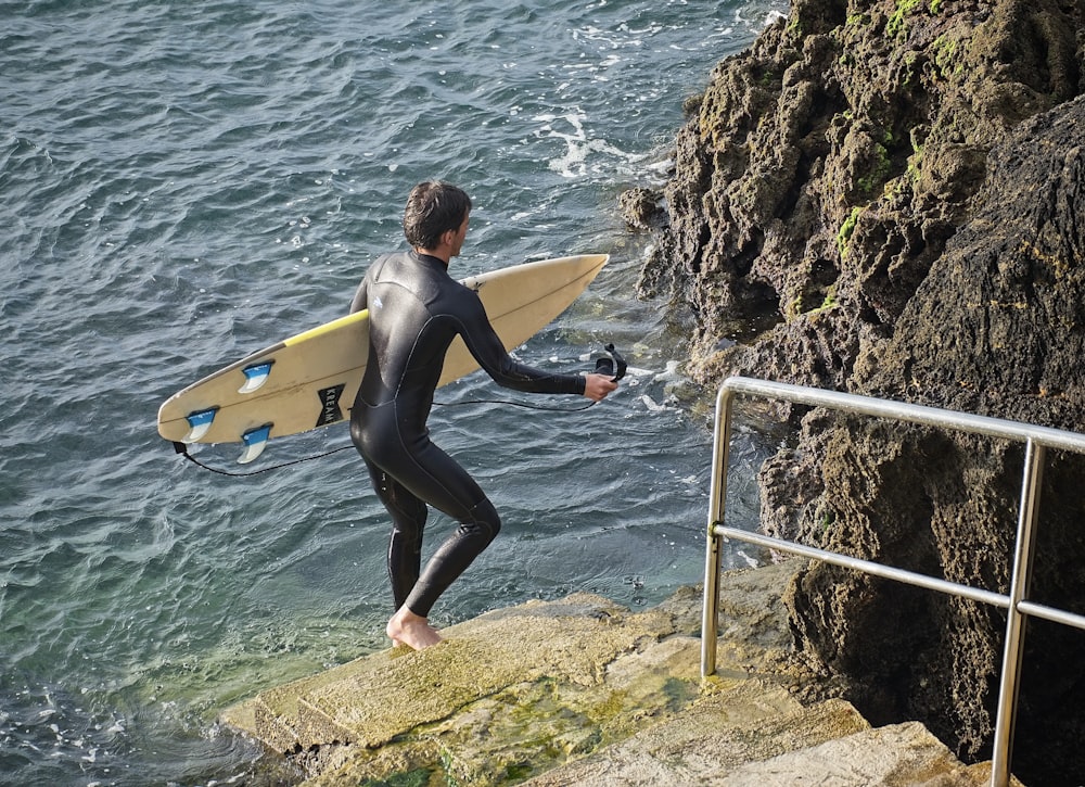 woman in black wetsuit holding black surfboard standing on rock near body of water during daytime