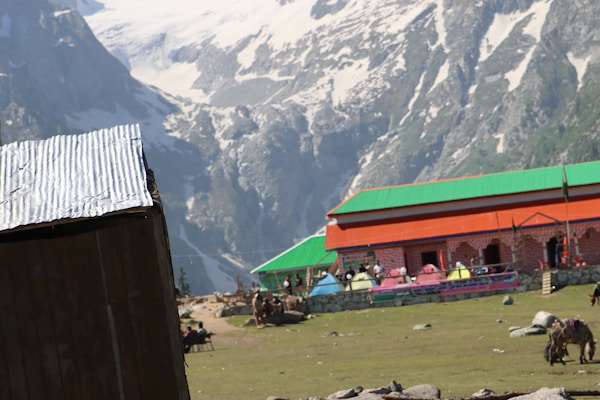 A building with a green roof and brick walls stands amidst a mountainous landscape. Snowy peaks rise in the background, and a corrugated metal structure is partially visible to the left. The foreground features a grassy area with several tents and grazing animals.