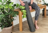 A person wearing gray patterned leggings is sitting on a wooden bench. They are holding a rolled-up yoga mat and are surrounded by potted green plants. The floor has a light wood texture, and there is a small statue on the bench.