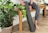A person wearing gray patterned leggings is sitting on a wooden bench. They are holding a rolled-up yoga mat and are surrounded by potted green plants. The floor has a light wood texture, and there is a small statue on the bench.
