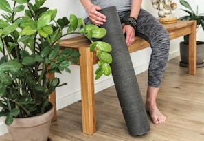 A person wearing gray patterned leggings is sitting on a wooden bench. They are holding a rolled-up yoga mat and are surrounded by potted green plants. The floor has a light wood texture, and there is a small statue on the bench.
