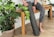 A person wearing gray patterned leggings is sitting on a wooden bench. They are holding a rolled-up yoga mat and are surrounded by potted green plants. The floor has a light wood texture, and there is a small statue on the bench.