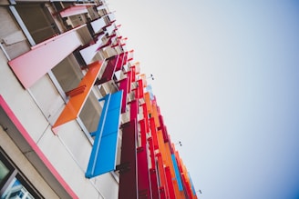 Vibrant and colorful panels in shades of pink, orange, red, and blue are attached to the exterior of a building, reaching upwards into the sky. These panels are positioned in a staggered pattern, creating a dynamic visual effect against the neutral tones of the building facade.