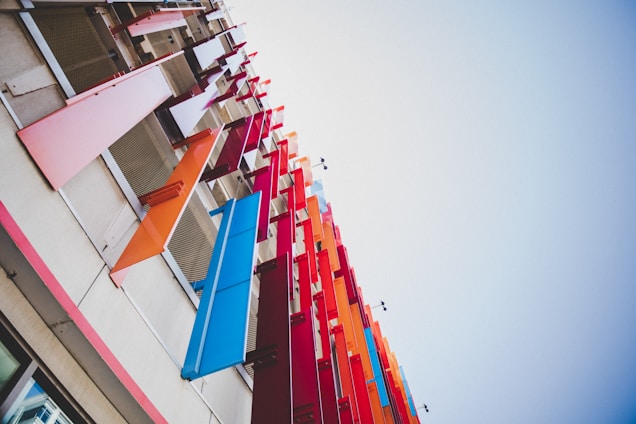Vibrant and colorful panels in shades of pink, orange, red, and blue are attached to the exterior of a building, reaching upwards into the sky. These panels are positioned in a staggered pattern, creating a dynamic visual effect against the neutral tones of the building facade.